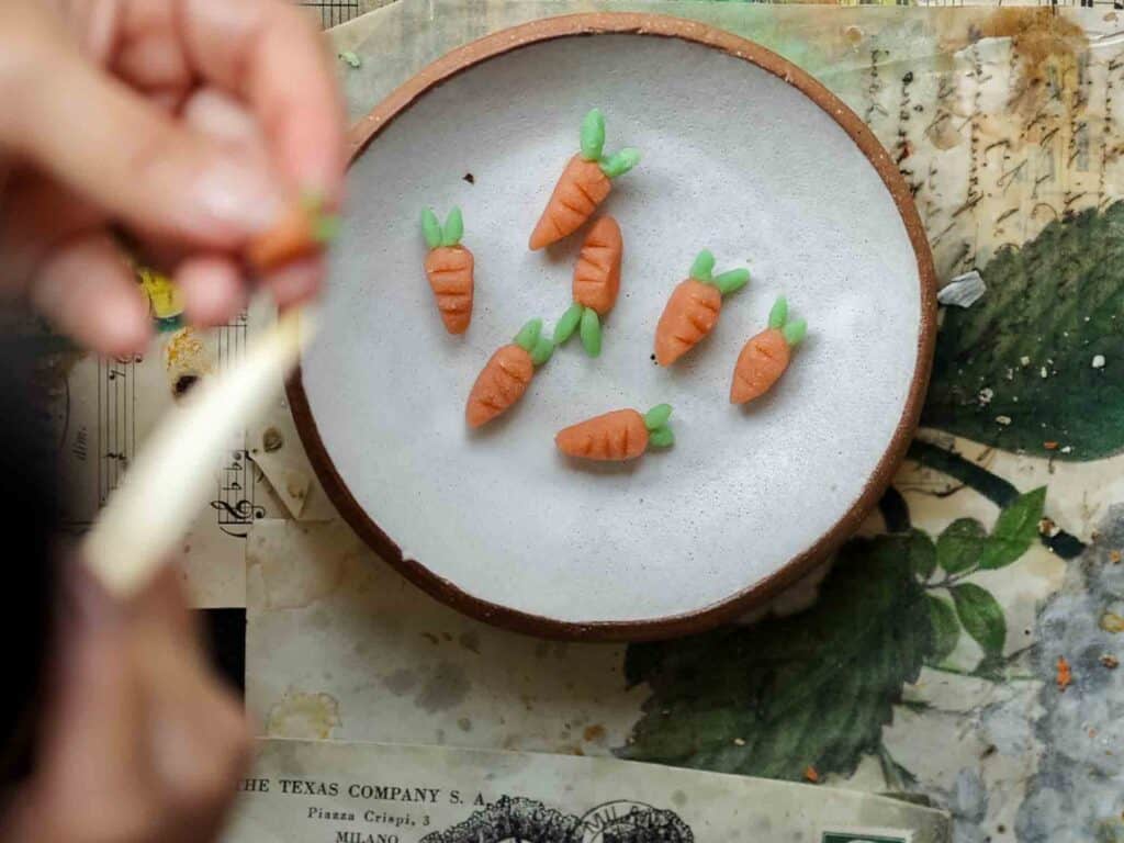 A person shaping small sculptures of carrots from marzipan or a similar material. Several finished carrot sculptures are placed on a round plate. The scene is set on a table with scattered papers and artistic supplies.